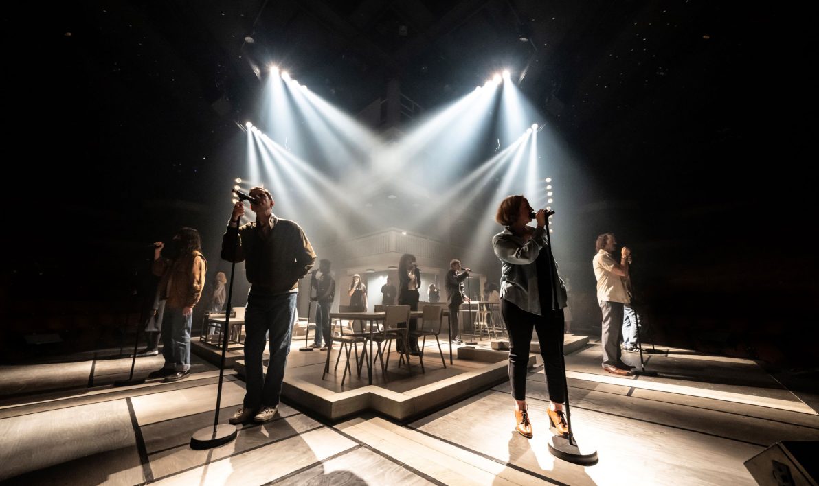 Cast of Standing at the Sky's Edge on stage at the National Theatre, London. The stage is dark, lit only by spotlights. The cast stand individually singing into microphones on stands.