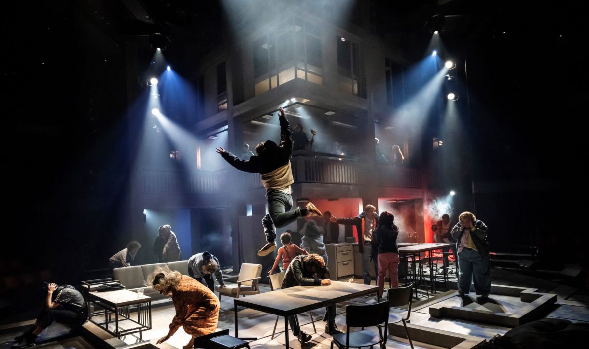 Cast of Standing at the Sky's Edge on stage at National Theatre, London. The stage is lit in spotlights, the cast dance around / on tables. One person jumping through the air in the centre of the image.